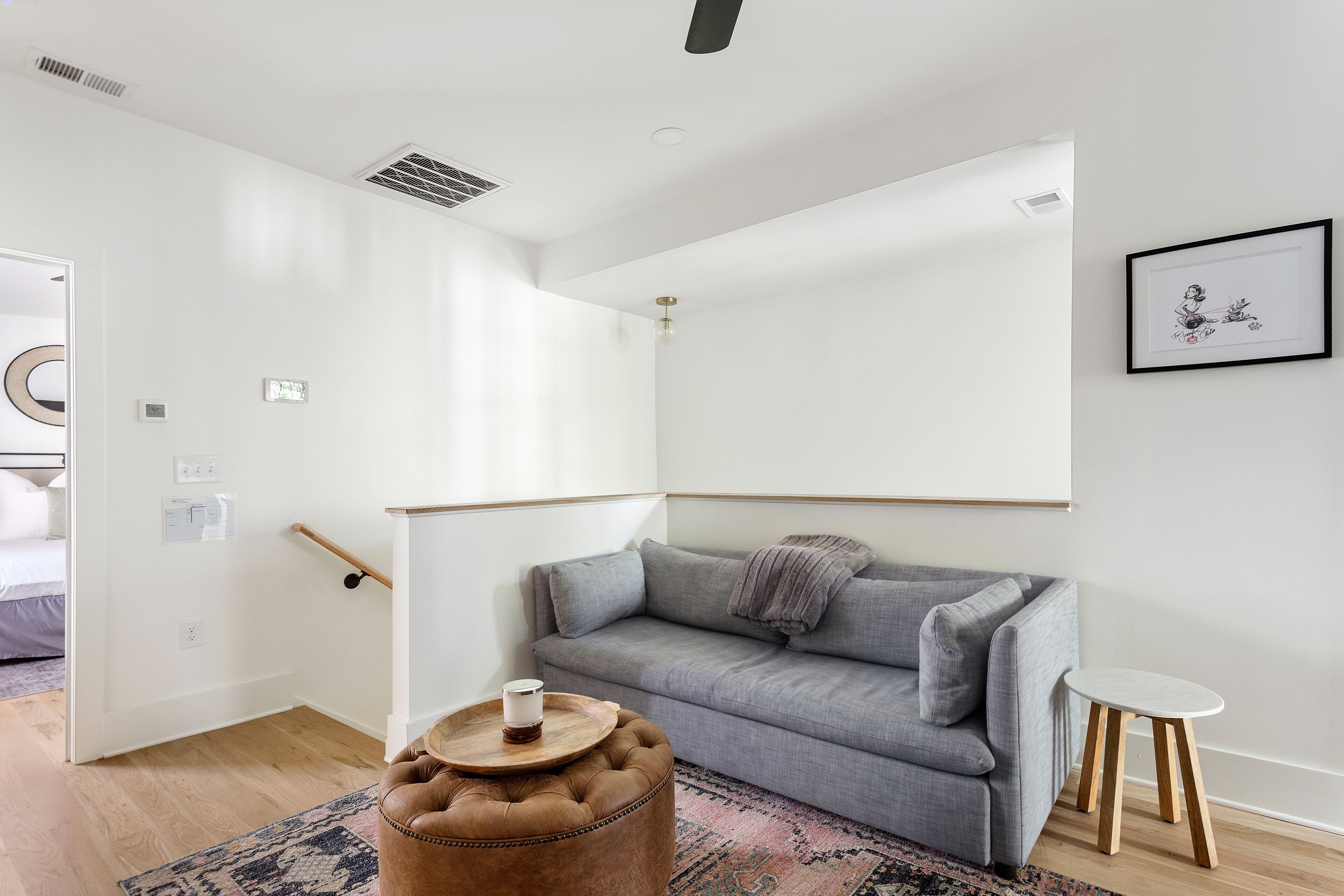 Ein modernes Wohnzimmer mit grauem Sofa, einem runden Hocker mit Kerze, einem kleinen weißen Tisch und einem gerahmten Bild an der Wand.