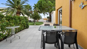 A patio area with a glass table and chairs, surrounded by greenery and a yellow wall.