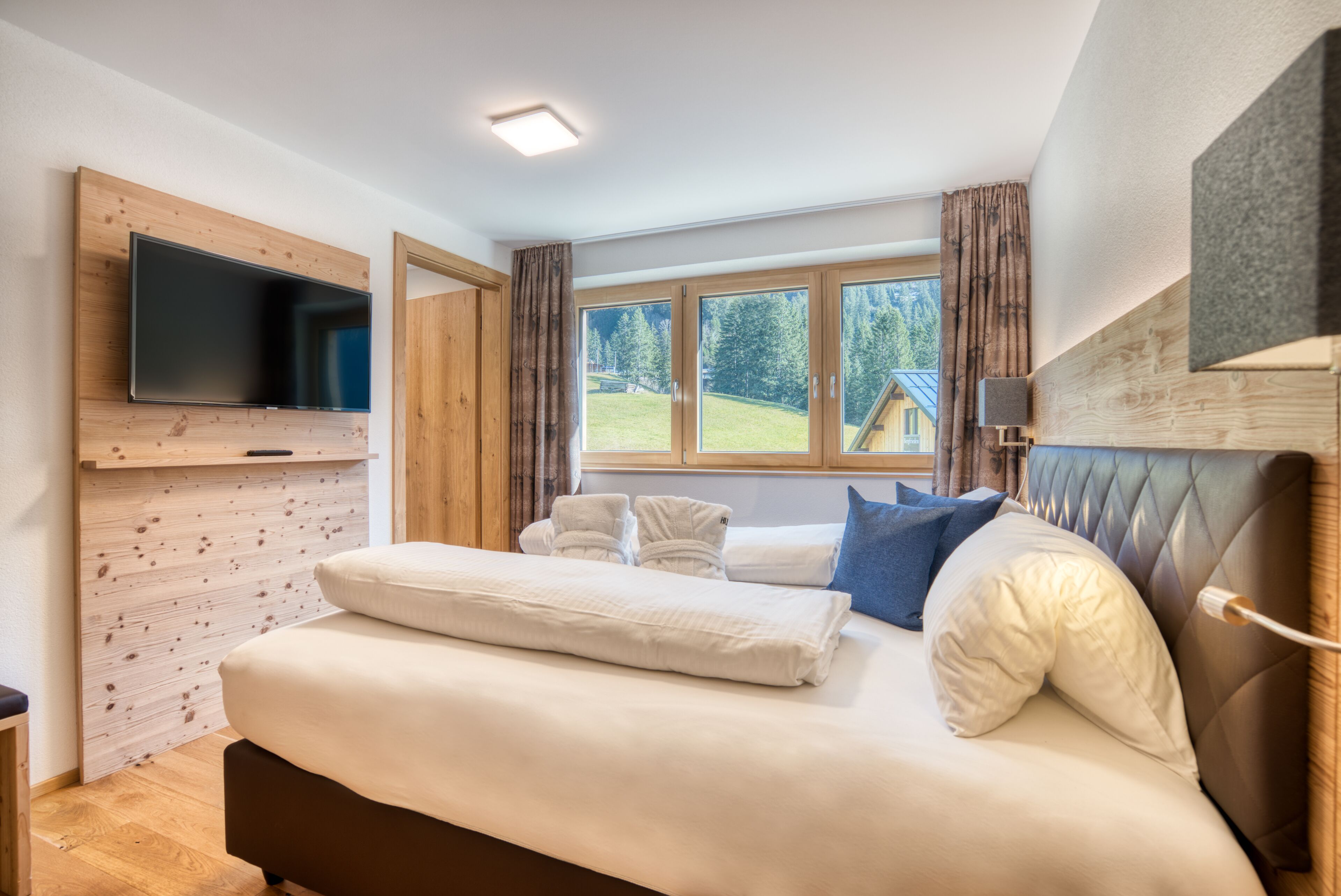 A bedroom with a large bed, a flat-screen TV mounted on the wall, and a window with curtains.