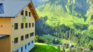 A multi-story building with a sign reading "HUS3 Apartments." It features a sloped roof and multiple windows. The surrounding landscape includes green hills, a valley, and a church spire. - HUS 23 (Schroecken)