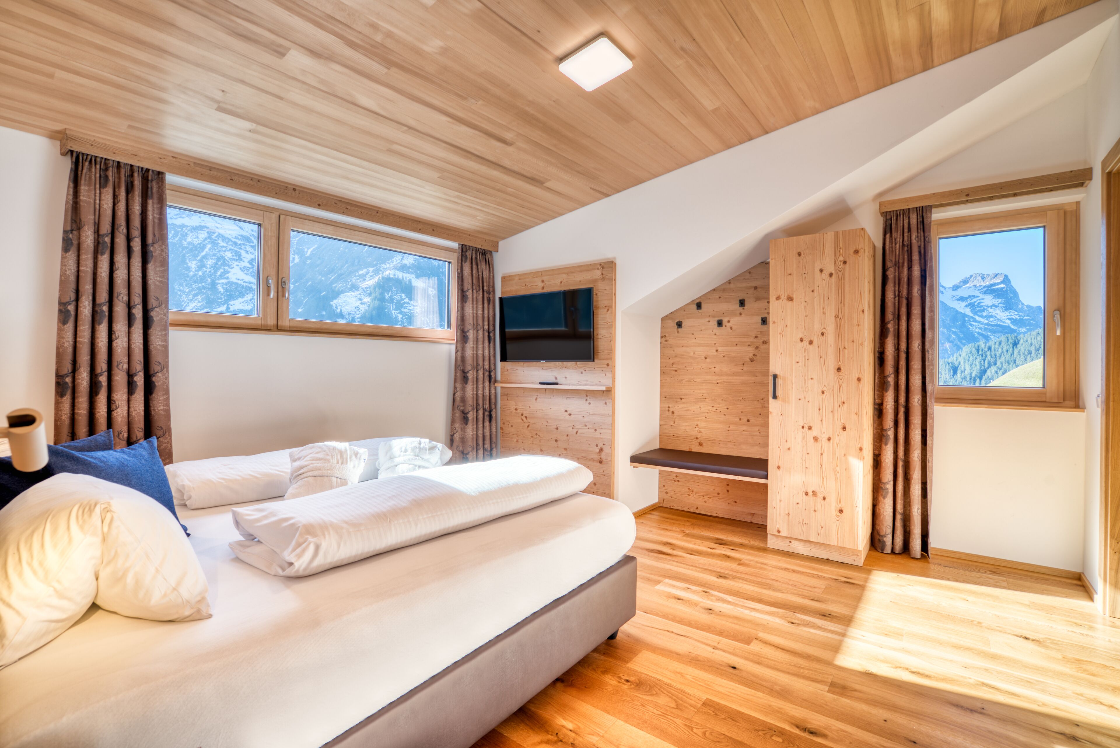 Een moderne slaapkamer met een groot bed, houten vloeren, een aan de muur bevestigde televisie en uitzicht op bergen door de ramen.