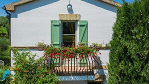 Un bâtiment blanc avec des volets verts, un petit balcon orné de fleurs rouges et un ciel d’un bleu limpide.