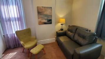 Un salon avec un fauteuil jaune, un canapé marron, une petite table avec une lampe et un tableau accroché au mur.