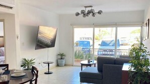 A spacious living room with a sofa, a dining table with chairs, a flat-screen TV mounted on the wall, and a large window with a view of the outside. - COMFORTABLE APARTMENT WITH POOL, WHIRLPOOL BATHTUB, 70" TV AND MORE (Cabo San Lucas)