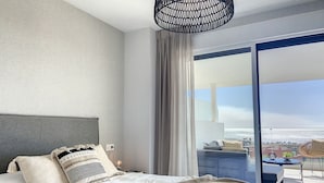 A bedroom with a bed, a nightstand, a lamp, and a view of the city through a large window. - Via Celere 2331 Golf & Sea view (Casares)
