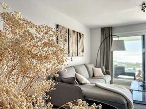 A modern living room with a grey sofa, a large window with curtains, and a decorative plant in the foreground.