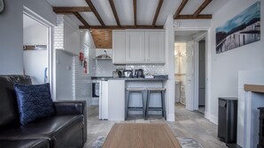 A modern kitchen with white cabinets, a central island with stools, and a dining area with a black leather sofa and patterned cushions. - Honeysuckle Cottage - 1 Bedroom Cottage - Amroth (Amroth)
