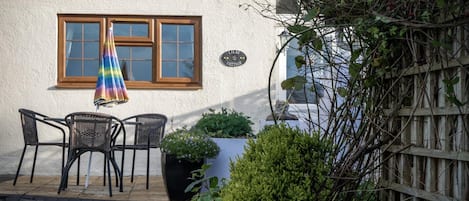 A cottage with a white exterior, a wooden window, and a sign reading "Lilac Cottage". There are two wicker chairs outside and a potted plant nearby.