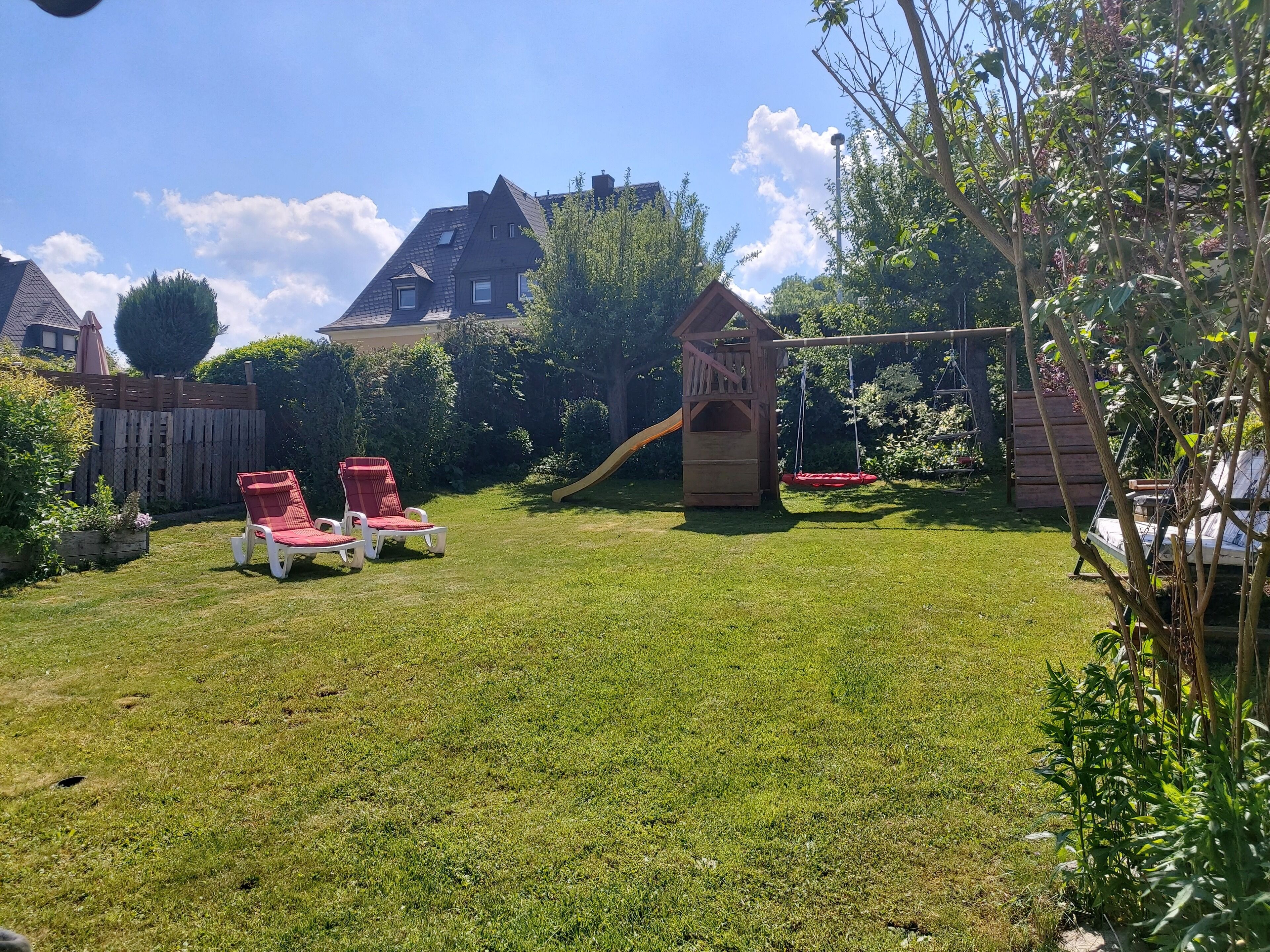 Un jardin avec une cabane en bois, un toboggan et deux fauteuils rouges.