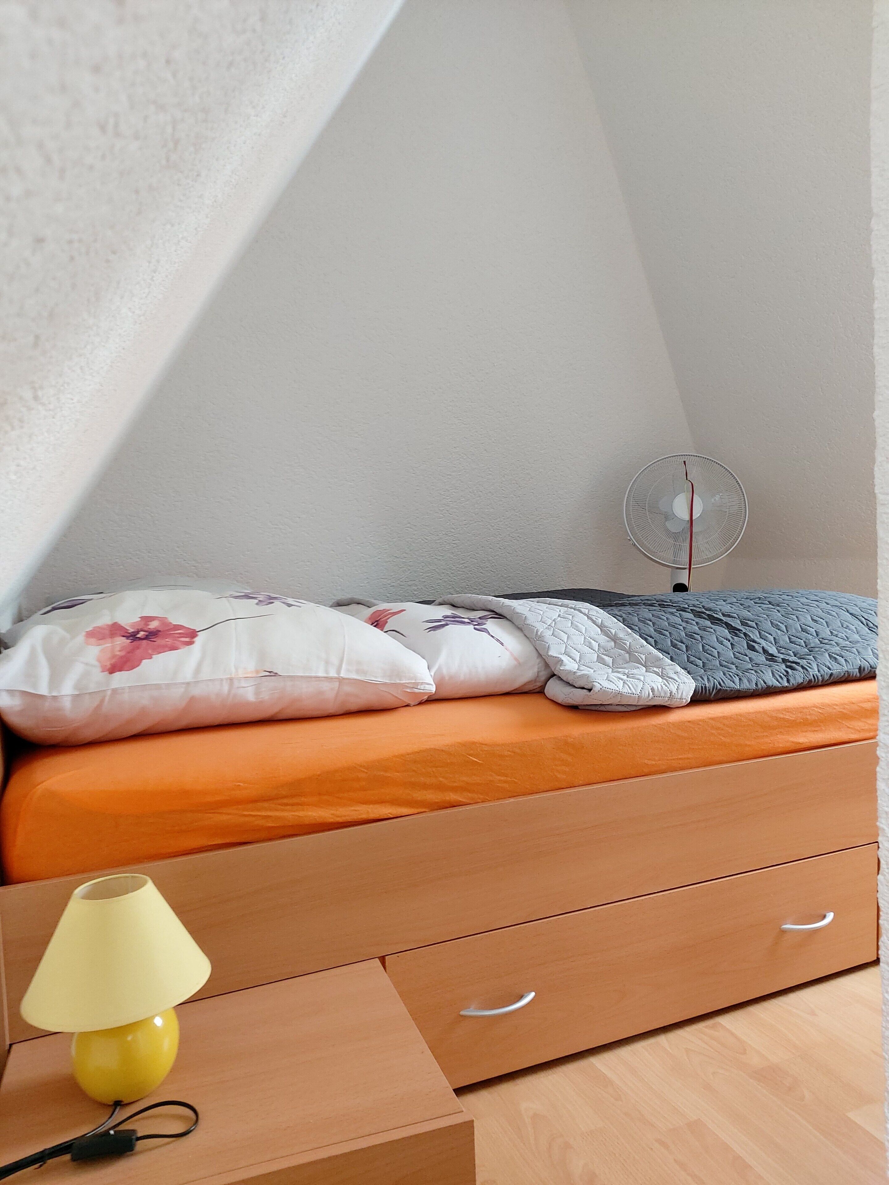 Une chambre à coucher avec un lit, des oreillers, une table de chevet avec une lampe, un ventilateur et une commode en bois.