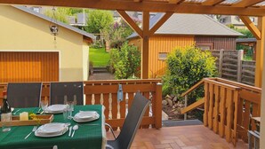 A covered outdoor dining area with a table set for two, a wooden pergola overhead, and a view of a garden and neighboring buildings.