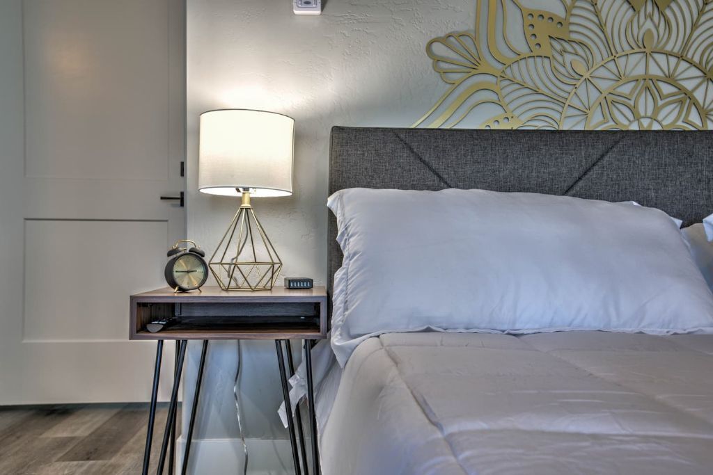 A neatly made bed with white linens, a grey upholstered headboard, and a bedside table with a lamp, alarm clock, and a geometric decorative piece.