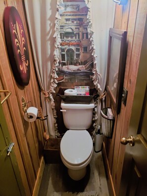 A small, narrow bathroom with a toilet, a roll of toilet paper, and a trash can. - The Loft at Old Falls (Lyles)