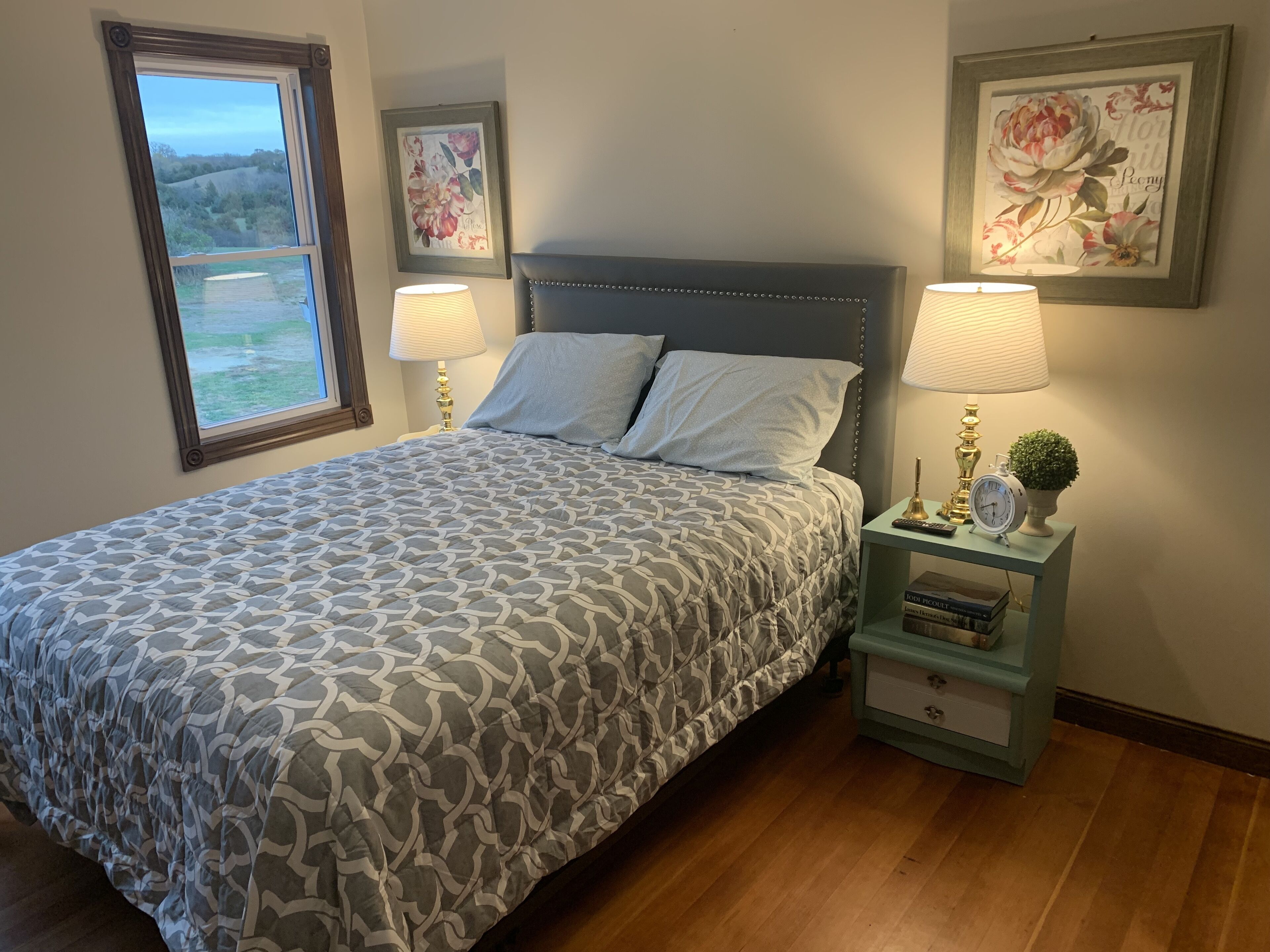 A neatly made bed with patterned bedding, two bedside lamps, a small bedside table with a clock and a plant, and framed floral artwork on the walls.