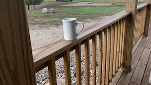 Una terraza de madera con una taza blanca en la barandilla, con vistas a un paisaje rural con campos, árboles y una masa de agua distante.