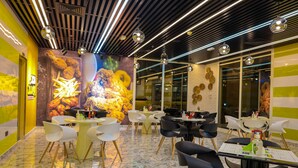 Un restaurante con un interior moderno, que presenta una combinación de mesas redondas y cuadradas, sillas blancas y negras, y un mural vibrante en la pared.