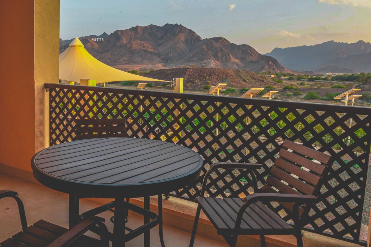 A balcony with a round table and two chairs, a yellow shade, and a view of mountains.