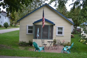 Relax on the porch with Adirondack chairs and a small table, surrounded by a serene lawn and trees.