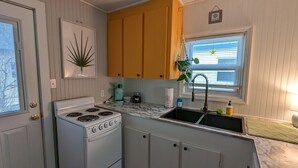 Kompakti keittiö, jossa on valkoinen liesi, keltaiset kaapit, pesuallas ja ikkuna, jossa on sälekaihtimet.