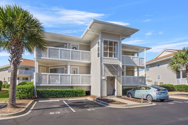 A multi-story building with balconies, a parking lot, and a car parked in front.