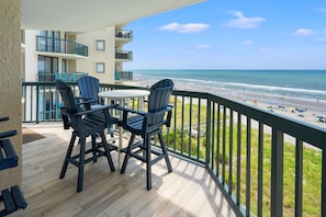 A balcony with two Adirondack chairs and a small table, overlooking a beach with people and umbrellas.
