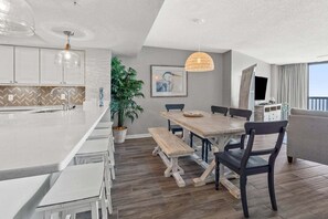 A modern kitchen with a central island, dining area with wooden chairs, and a large window with curtains. - Light & breezy oceanfront condo rental with lazy river, pool, and fitness center near Ocean Drive (North Myrtle Beach)