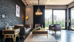 Una sala moderna con un sofá de cuero, una mesa de centro de vidrio y un sillón de cuero. Se observa una pared de ladrillo negro, un ventanal grande y una lámpara colgante.
