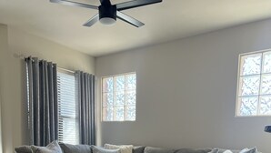 A living room with a sectional sofa, a ceiling fan, and a large window with blinds.