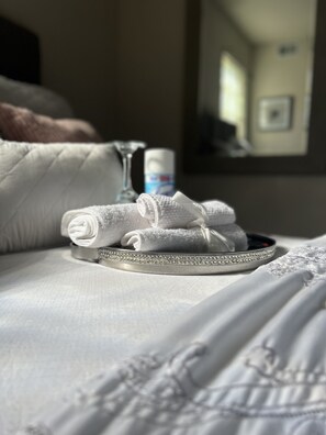 Un lit bien fait, avec du linge de lit blanc, un plateau contenant des serviettes pliées, un verre et une bouteille de lotion.