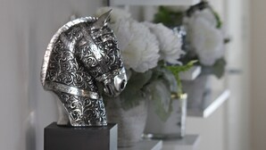 A silver decorative horse head sculpture on a white shelf, surrounded by white flowers and books.