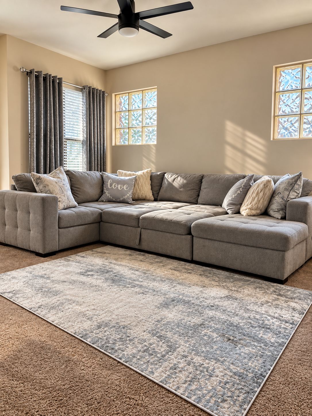 A sectional sofa with multiple cushions, a ceiling fan, and a patterned rug in a room with windows and curtains.