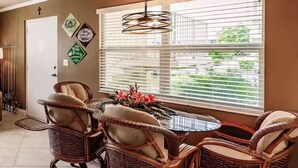 A dining area with wicker chairs and a round table, a window with blinds, and wall decorations. - Margaritaville, Steps From The Snook Inn, Olde Marco (Marco Island)
