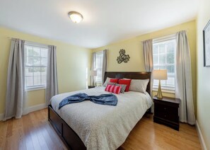 Un dormitorio con una cama, dos mesitas de noche, una lámpara de pared y una ventana con cortinas.