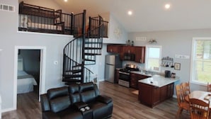 Un vaste espace de vie comprenant un canapé en cuir noir, une table à manger avec des chaises, une cuisine dotée d’un îlot central et un escalier en colimaçon menant à une chambre en mezzanine.