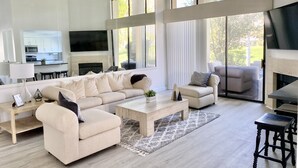 Ein großzügiger Wohnraum mit einer großen Couch, einem Couchtisch und einem Kamin. Durch einen offenen Durchgang ist eine Küche erkennbar, und große Fenster sorgen für natürliches Licht.