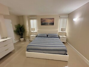 A bedroom with a bed, bedside tables, a nightstand, a potted plant, and a painting on the wall.