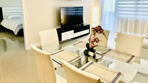 A modern hotel room with a glass dining table, white chairs, a flat-screen TV, a minibar, and a large window with sheer curtains.