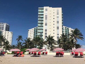 A beachfront hotel with multiple floors, palm trees, and red and white striped beach umbrellas.