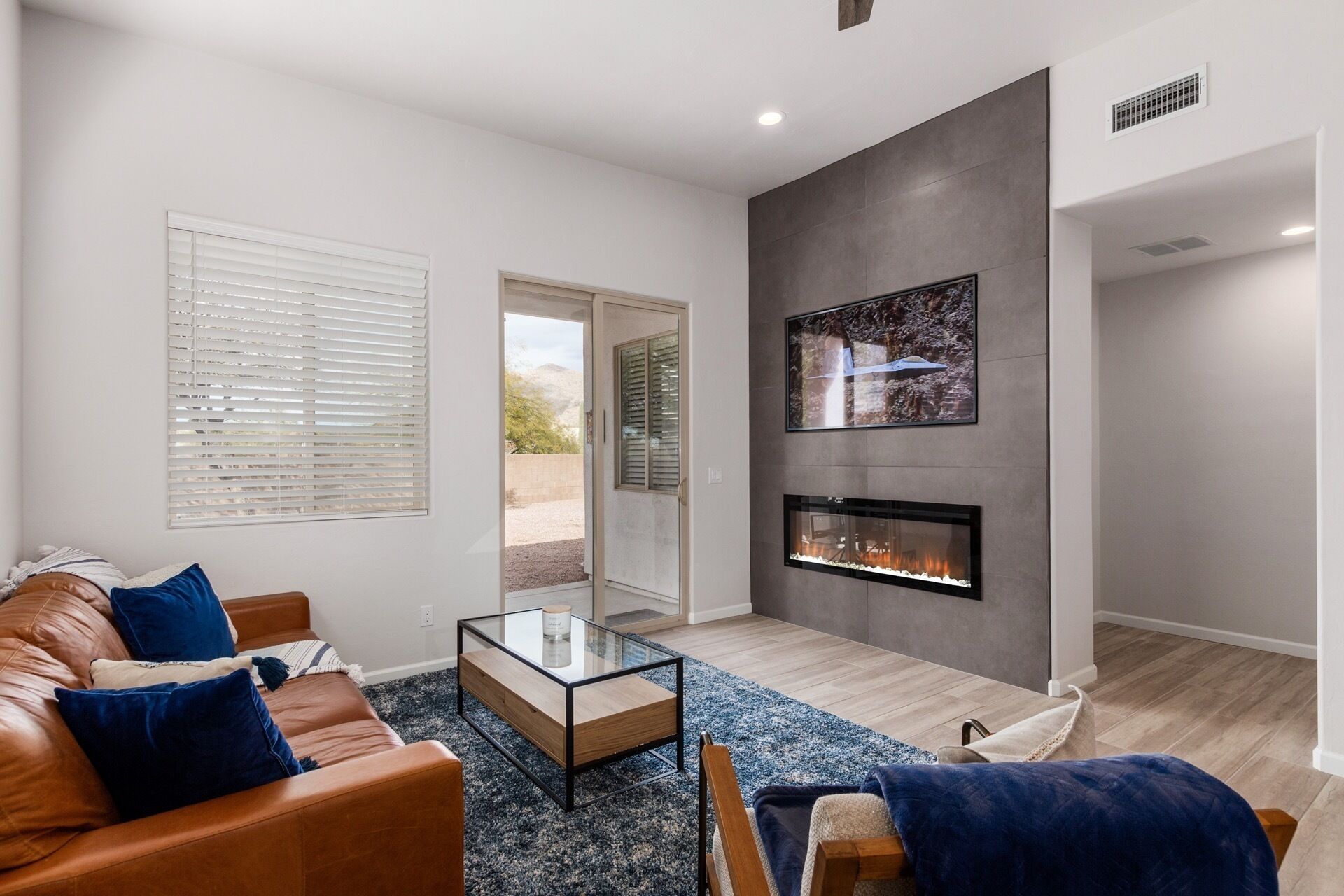 A modern living room with a leather sofa, a glass coffee table, a fireplace, and a large window with blinds.