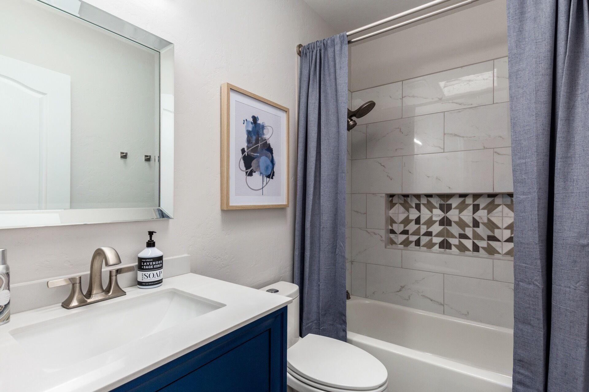 Ein Badezimmer mit einem weißen Waschbecken, einem blauen Schrank, einem gerahmten abstrakten Gemälde, einer Dusche mit geometrischem Fliesendesign und einer weißen Toilette.