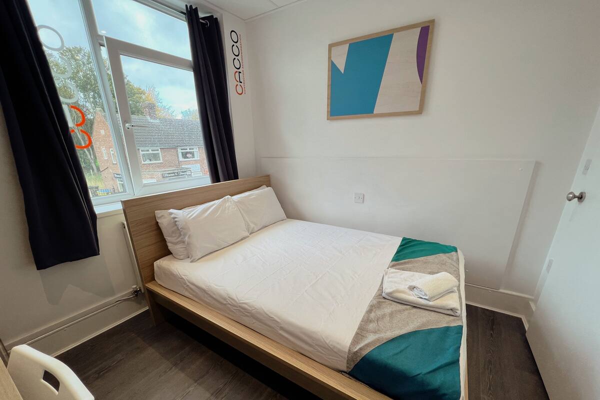 A single bed with a wooden frame, white bedding, and a teal and beige throw blanket. A window with curtains is visible, and a wall-mounted artwork is present.