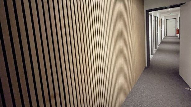 A corridor with a wooden slat wall, a grey carpeted floor, and a white door at the end. - Contractor Accommodation - Cacco (Corby)