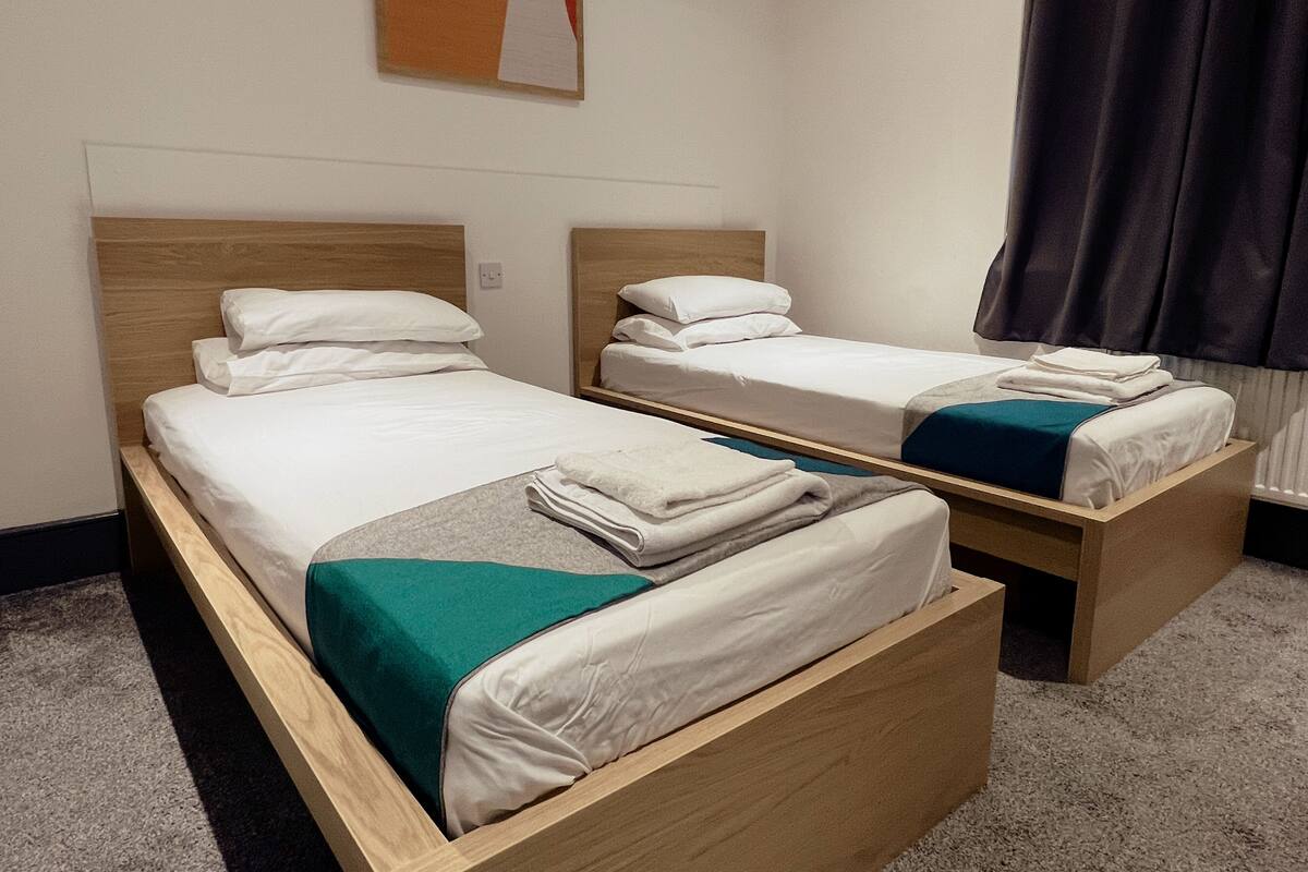 Two single beds with wooden frames and white bedding, a grey carpet, and a dark curtain on a window.