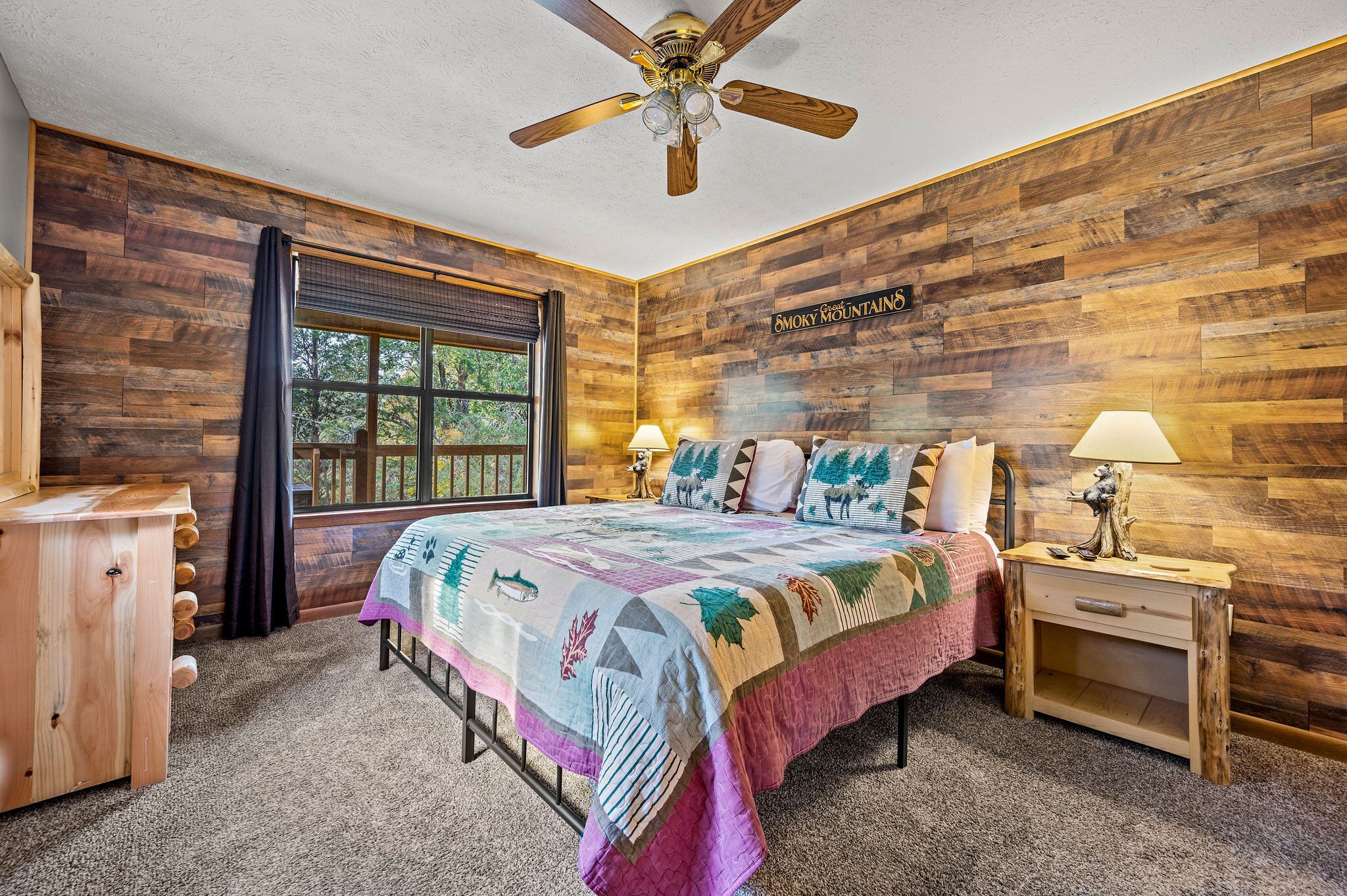 A bedroom with a wooden wall, a bed with a colorful quilt, a ceiling fan, and a window with a view of trees.
