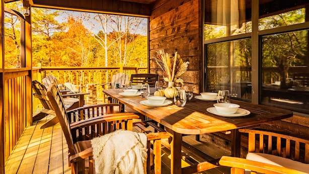 A wooden deck with a dining table set for four, surrounded by chairs, and large windows offering a view of autumn trees. - Family ‑Friendly Retreat: Hot Tub, Theater + Close to Attractions (Sevierville)