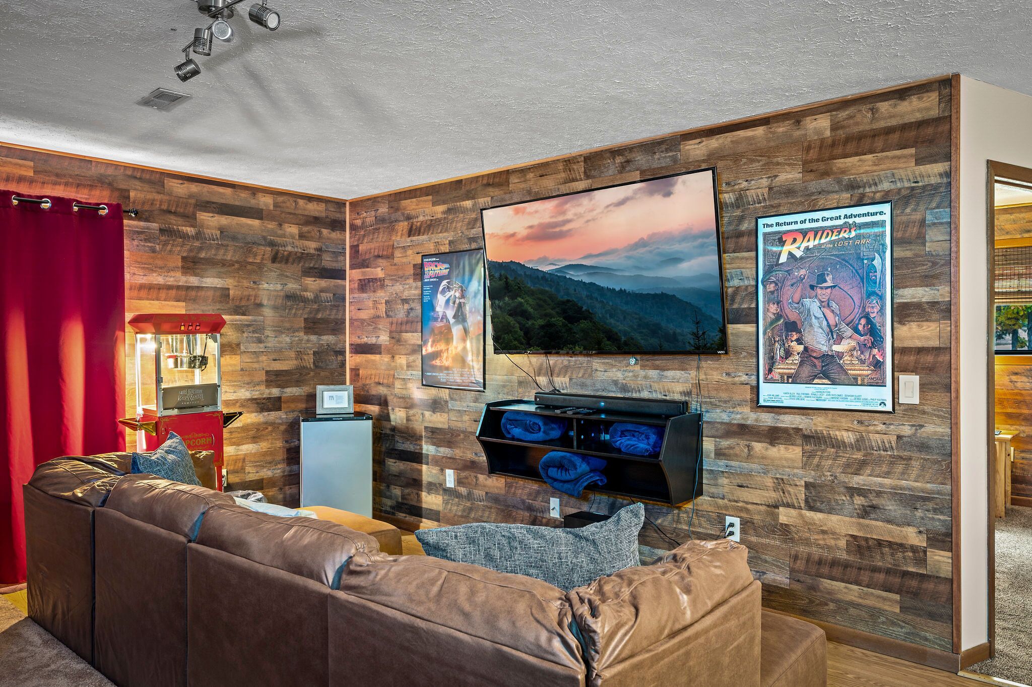 A living room with a wooden wall, a large flat-screen TV, a leather sofa, and a small table with a lamp.