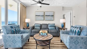 A room with a ceiling fan, two framed artworks, a grey sofa with patterned cushions, a round wooden coffee table, and two lamps on side tables.