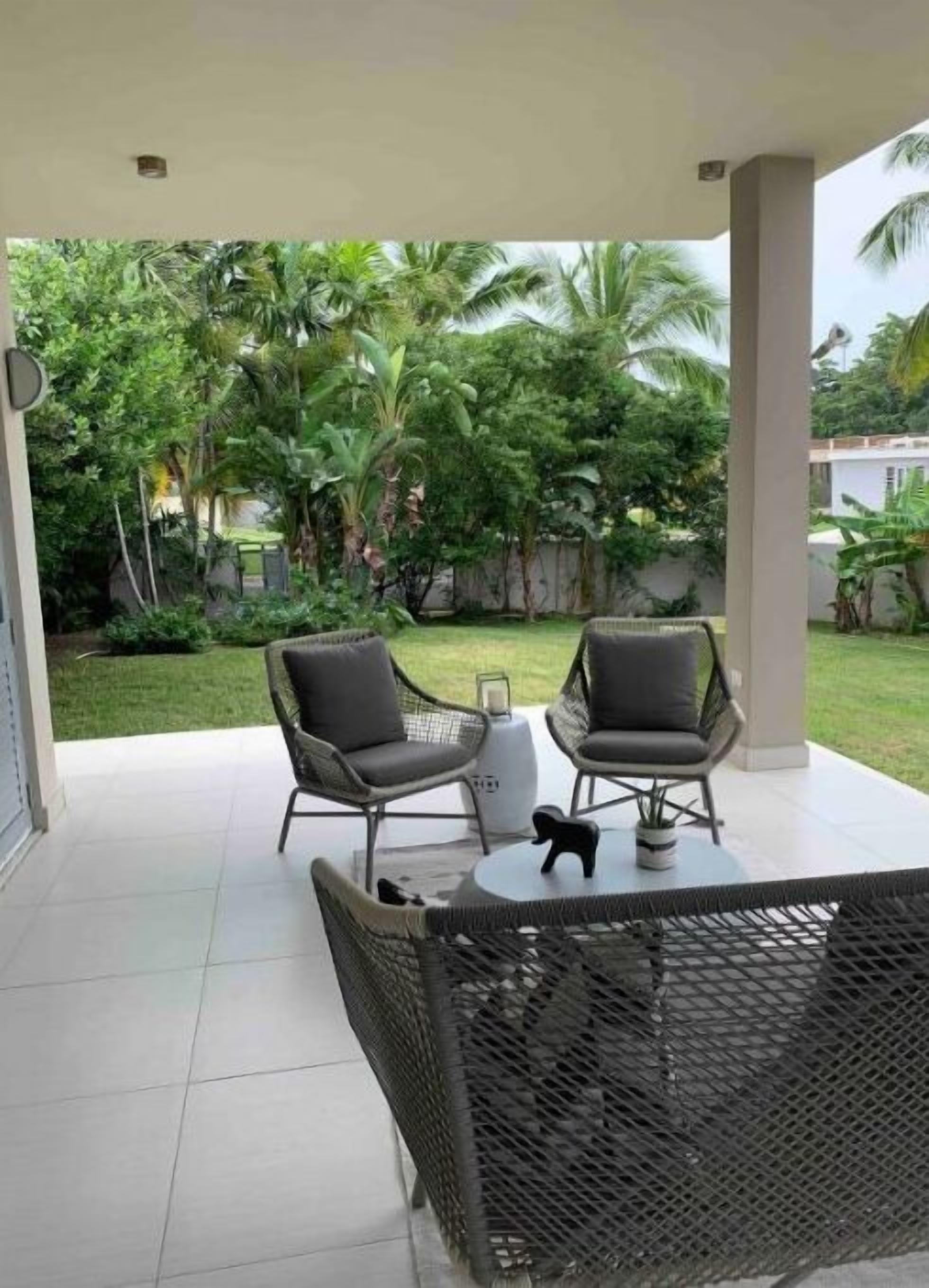 Ein überdachter Patio-Bereich mit zwei grauen Stühlen und einem Tisch, umgeben von üppiger Bepflanzung und Palmen.