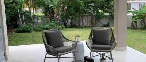 Ein überdachter Patio-Bereich mit zwei grauen Stühlen und einem Tisch, umgeben von üppiger Bepflanzung und Palmen.