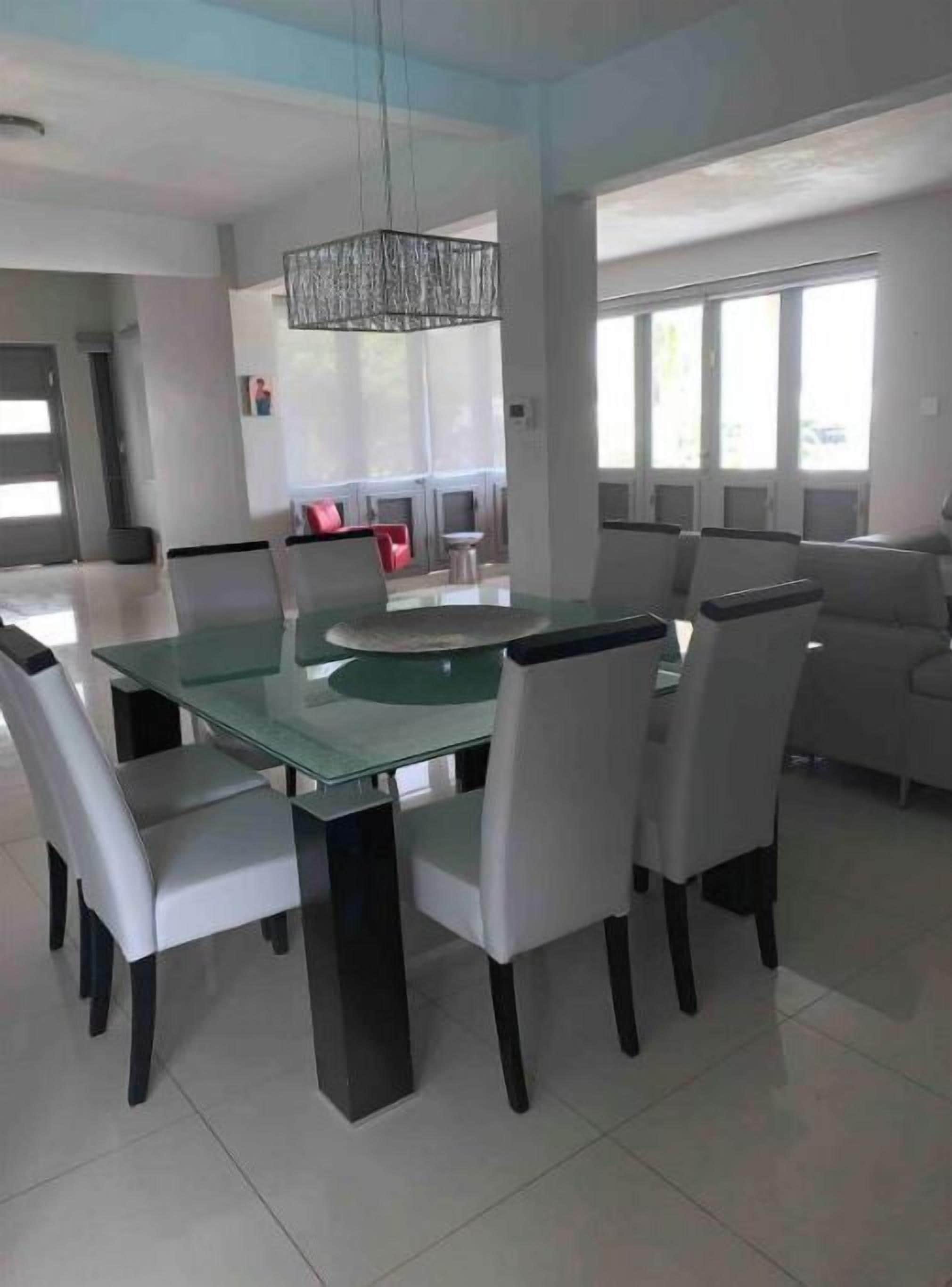 Uma área de refeições moderna com mesa de vidro e cadeiras brancas, um lustre e vista para uma sala de estar com sofá e lareira.